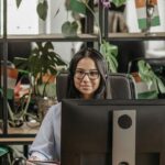 Asian woman with eyeglasses working at a desk in a modern office, surrounded by plants.
